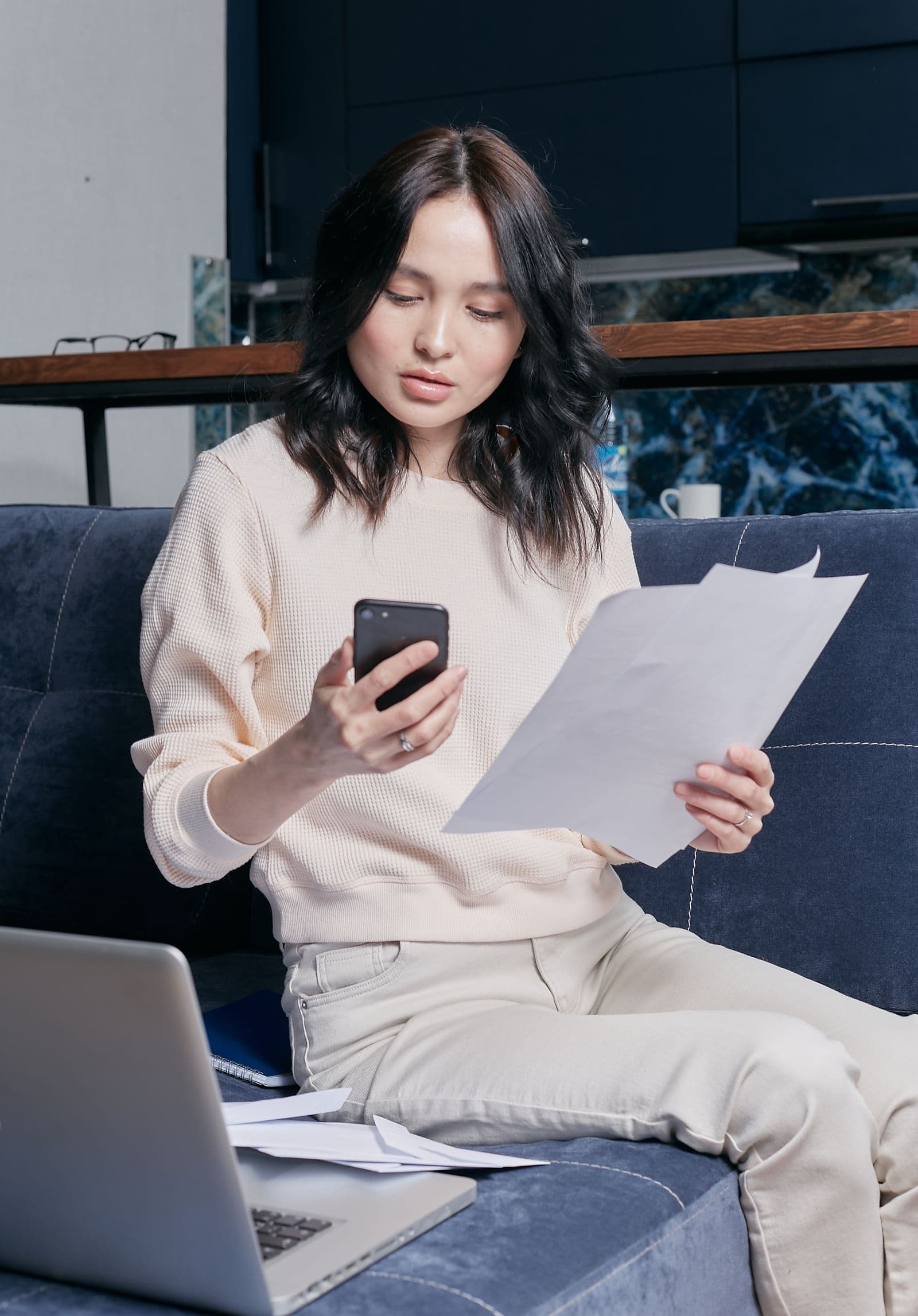 A woman checking documents with a mobile phone beside a laptop