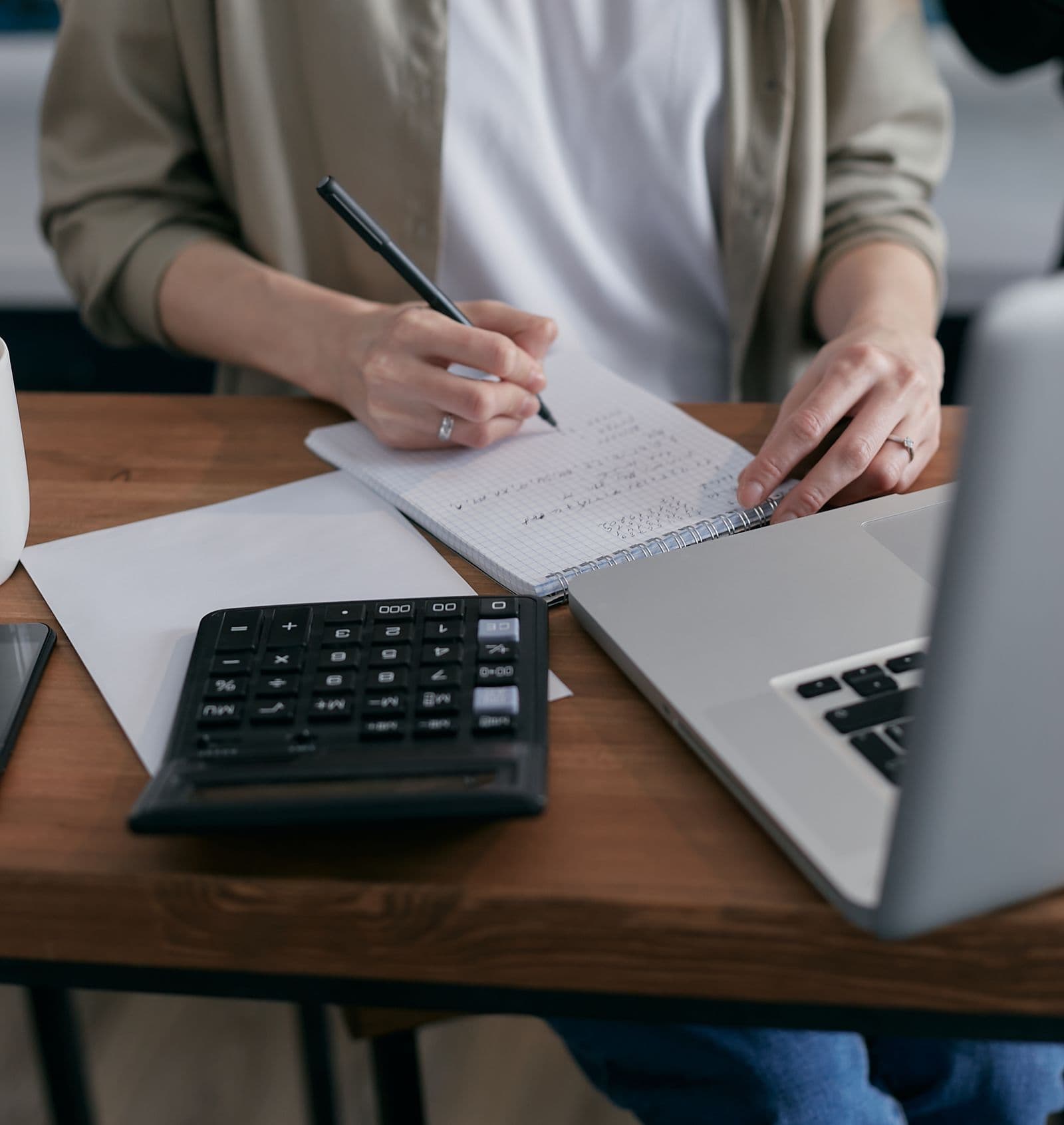 Hands writing calculations beside a calculator and laptop